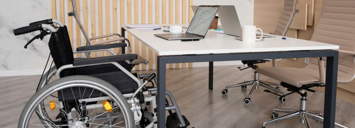 mesa de ambiente de trabalho com tampo branco e pés na cor cinza, com cadeira de rodas posicionada do lado esquerdo e uma cadeira convencional, com encosto e assento estofado na cor bege, à frente, do lado direito.