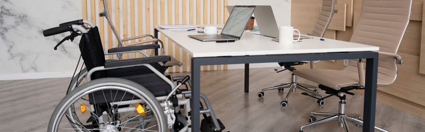 mesa de ambiente de trabalho com tampo branco e pés na cor cinza, com cadeira de rodas posicionada do lado esquerdo e uma cadeira convencional, com encosto e assento estofado na cor bege, à frente, do lado direito.