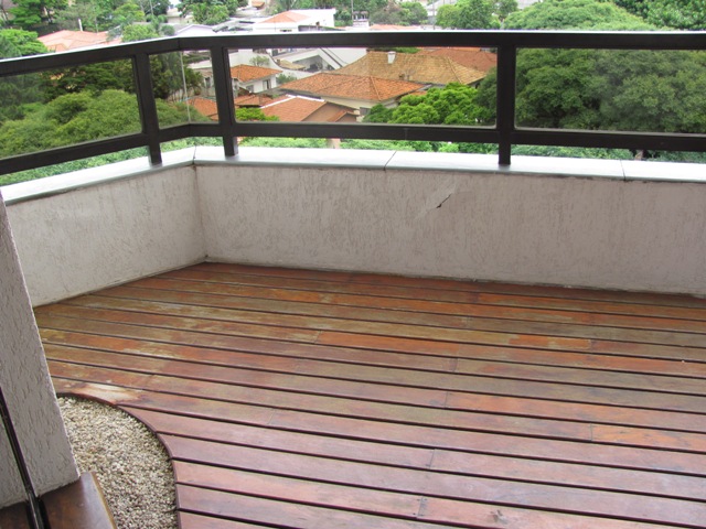 Vista da varanda antes da intervenção, com piso de madeira bem danificado, mureta branca e guarda-corpo de alumínio com vidro.