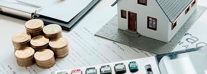 vista superior de parte de itens sobre superfície plana clara, sendo uma calculadora, a maquete de uma casa, sete pilhas de moedas em alturas diferente e um caderno.