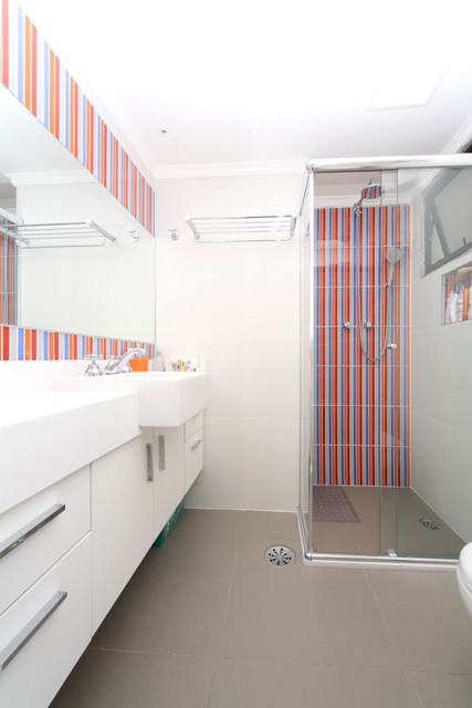 Vista no banheiro da suíte após a reforma, com piso na cor cinza, móveis e revestimentos brancos, com detalhes em filetes em vidro coloridos emoldurando o espelho e em uma parede do box.