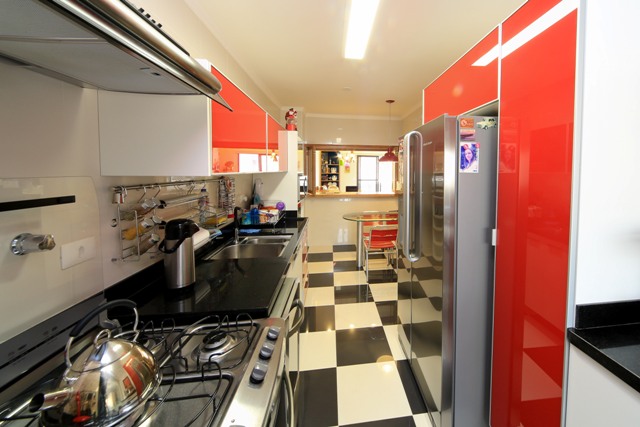 Vista da cozinha, após a reforma, a parti da lavanderia, mostrando o mobiliários com portas de vidro vermelho, nova bancada em granito preto e piso quadriculado preto e branco.