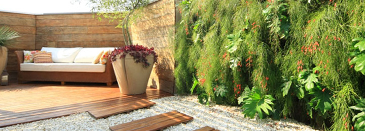 Área externa após intervenção, com piso em deck de madeira ao fundo, junto a um sofá de vime com estofado branco e dois vasos grandes aos lado, e pedriscos em seguida, onde encontra-se um muro verde.