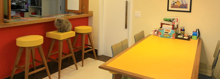 sala residencial, à esquerda um balcão que separa a sala da cozinha, com tampo de madeira e azulejos decorados, a parede frontal pintada de vermelho, com três banquetas alta à frente, com pés ed madeira,estofados amarelos e com um gato cinza sobre a do meio. À direita, uma mesa em madeira com tampo retangular amarelo, borda de madeira clara e quatro cadeiras na cor cinza.