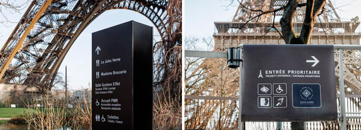 Duas imagens dispostas lado a lado de duas placas de sinalização da Torre Eiffel, ambas na cor preta, escrita em branco e com a torre ao fundo. As duas placas mostram, em ângulos diferentes, sinalizações de acessibilidade para acessar o monumento.
