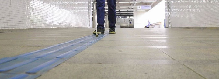 Vista baixa, da altura do piso, mostrando as pernas de um homem, vestindo calça jeans e tênis pretos, e caminhando de frente sobre uma faixa de piso direcional na cor azul, segurando uma bengala branca.