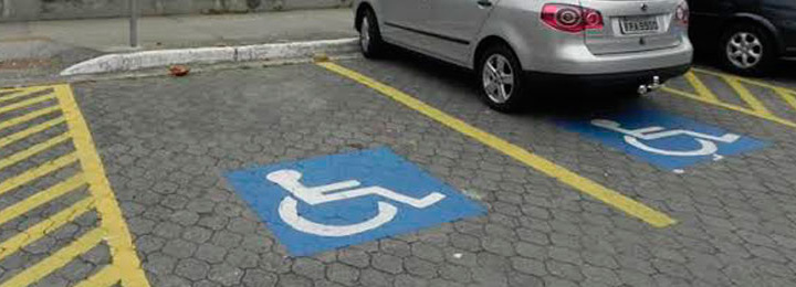 vagas reservadas para pessoa com deficiência pintadas no piso de um estacionamento em área externa, dispostas à 90 graus do meio fio, mostrando duas pinturas do símbolo internacional de acesso e com dois carros estacionados.
