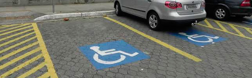 vagas reservadas para pessoa com deficiência pintadas no piso de um estacionamento em área externa, dispostas à 90 graus do meio fio, mostrando duas pinturas do símbolo internacional de acesso e com dois carros estacionados.